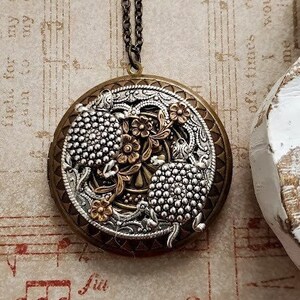 Pode incluir: Um colar com um medalhão redondo de prata e ouro, adornado com um design floral detalhado. O medalhão é redondo e tem uma abertura com dobradiça. A corrente é de prata e tem um padrão de elos delicados.