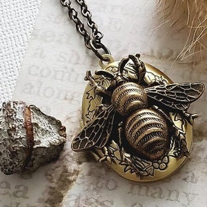 May include: A gold-toned locket necklace with a detailed bee design. The bee is surrounded by floral accents and has a textured, striped body. The necklace has a black chain.