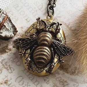May include: A gold-toned locket necklace featuring a detailed bee design. The bee is surrounded by floral accents and has a textured, striped body.