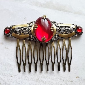 May include: A gold hair comb with a red glass cabochon in the center and two smaller red glass cabochons on either side. The comb has a decorative filigree design.