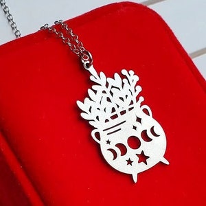 Puede incluir: Un collar con colgante plateado con un diseño de caldero adornado con lunas crecientes, estrellas y follaje. El collar se muestra sobre una superficie roja, resaltando los detalles de esta joya de temática de brujería.