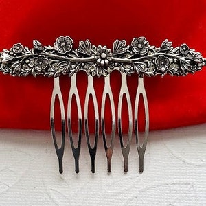 May include: Silver hair comb with a floral design. The comb has seven teeth and is decorated with leaves and flowers.