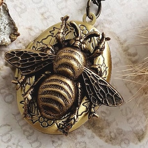 Puede incluir: Un collar dorado con un medallón que presenta un diseño de abeja detallado. El medallón es de forma ovalada y tiene una superficie texturizada. La abeja se representa con alas intrincadas y un cuerpo segmentado.