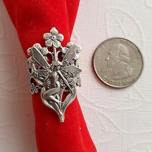 May include: Silver ring featuring a fairy with wings, sitting on a flower stem with two flowers. The ring is on a red velvet background.