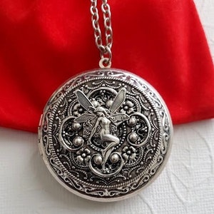 May include: A silver locket necklace with a detailed design featuring a fairy with wings. The locket is round and has a filigree pattern.