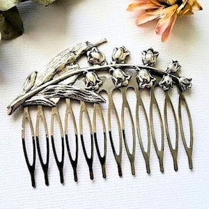 May include: Silver hair comb featuring a delicate lily of the valley design. The comb has a row of teeth and a floral motif with leaves and flowers.