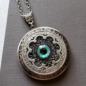 May include: Silver locket necklace with a detailed filigree design and a blue eye in the center.