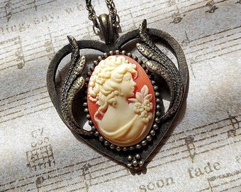 May include: A gold heart-shaped pendant necklace with a cameo of a woman's profile in a white cameo with a peach background. The cameo is surrounded by a beaded border.