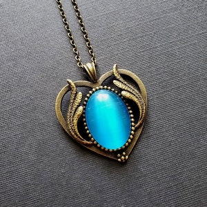 May include: A blue glass oval cabochon stone set in a gold-toned heart-shaped pendant with ornate details. The pendant is attached to a gold-toned chain.