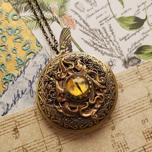 May include: A gold locket necklace with a detailed design of a dragon's eye with a yellow and green gemstone. The locket is surrounded by a floral design.