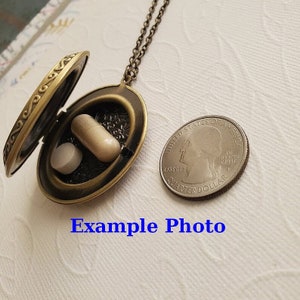 May include: A bronze locket necklace with a round, ornate design. The locket is open, revealing two small, white pills inside. A US quarter is shown for scale.