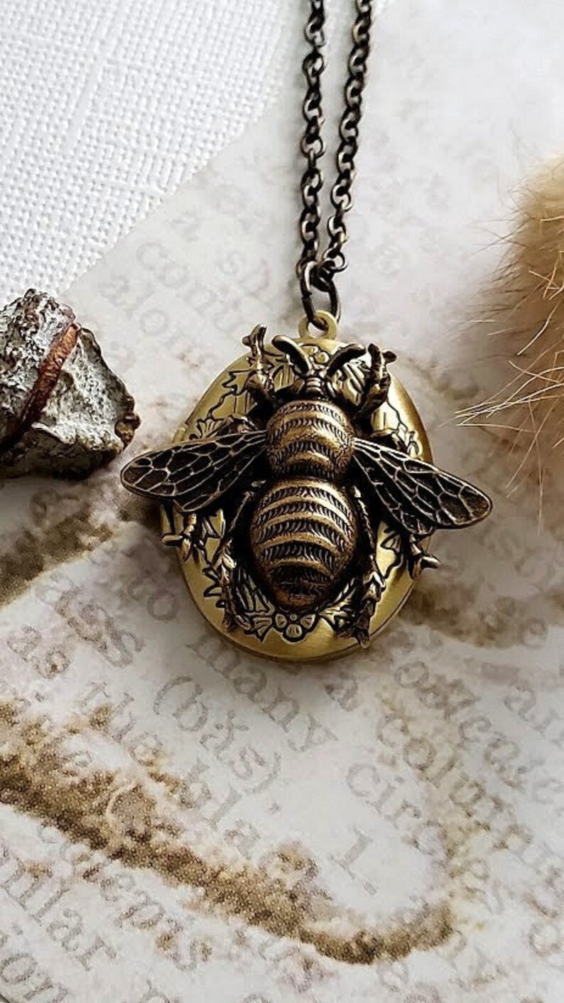 May include: A gold bee locket necklace with a detailed bee design and floral accents. The locket is oval shaped and hangs from a delicate chain.