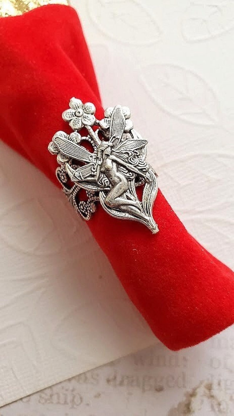 May include: Silver napkin ring featuring a fairy with wings and flowers. The fairy is sitting on a leaf with her wings spread out. The ring is ornate and detailed.