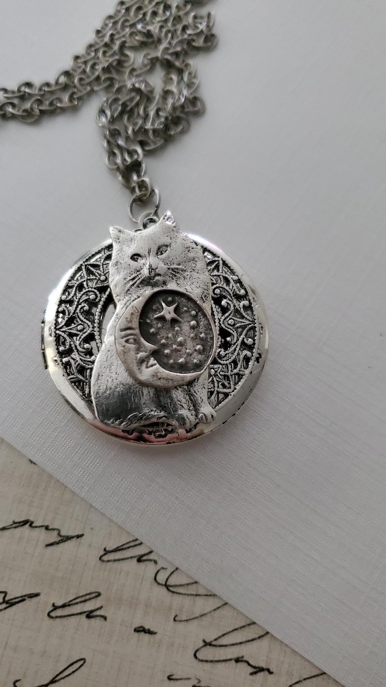 K&ouml;nnte beinhalten: Ein silberfarbener, runder Medaillon-Anh&auml;nger an einer passenden Kette. Das Medaillon zeigt ein erhabenes Relief einer Katze, die einen Halbmond mit einem Stern vor einem texturierten Hintergrund h&auml;lt. Der Rand hat ein filigranes Muster.