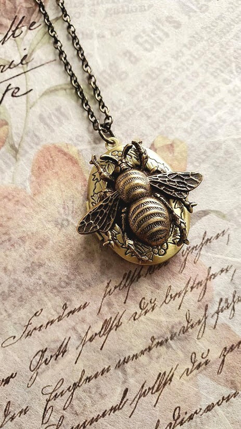 May include: A gold-toned locket necklace featuring a detailed bee design. The locket is oval-shaped and has a floral pattern around the bee.