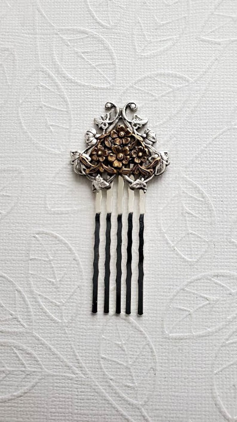 May include: Silver hair comb with a floral design. The comb has five teeth and a decorative top with a floral pattern in gold.