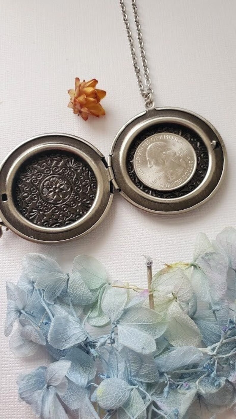 May include: A silver locket necklace with a detailed floral design on the front and a silver coin inside. The locket is open and the coin is visible. The necklace is on a white background.