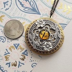 May include: A gold locket necklace with a detailed design featuring a dragonfly, flowers, and a yellow eye. The locket is about the size of a quarter.