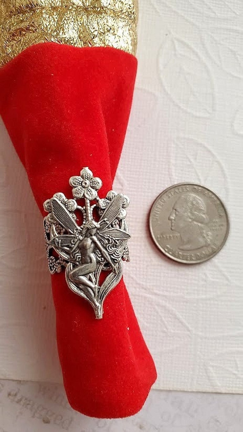 May include: Silver ring featuring a fairy with wings, sitting on a flower stem with two flowers. The ring is on a red velvet background.