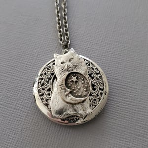 K&ouml;nnte beinhalten: Ein silberfarbener, runder Medaillon-Anh&auml;nger an einer passenden Kette. Das Medaillon zeigt ein erhabenes Relief einer Katze, die eine Mondsichel mit einem Stern und kleinen Punkten h&auml;lt. Der Hintergrund hat ein filigranes, durchbrochenes Muster.