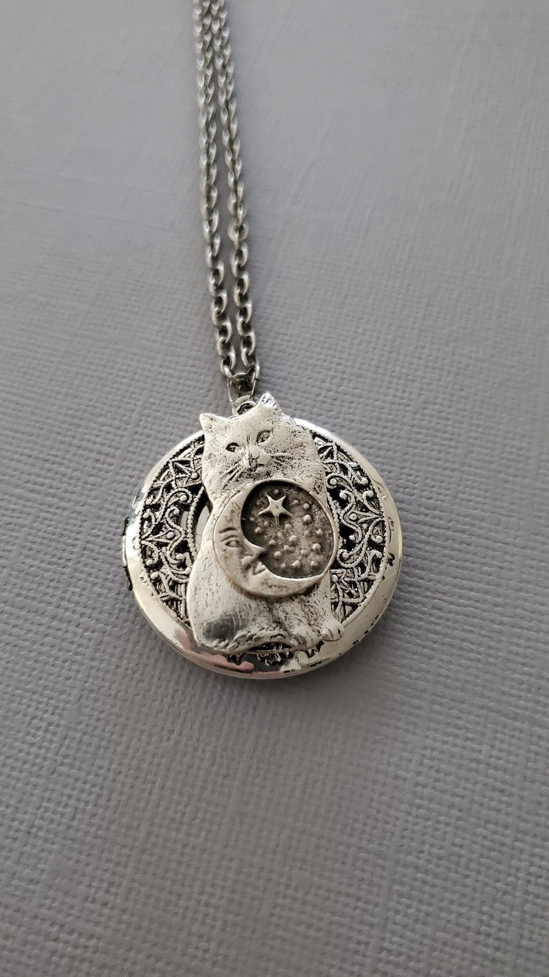 K&ouml;nnte beinhalten: Ein silberfarbener, runder Medaillon-Anh&auml;nger an einer passenden Kette. Das Medaillon zeigt ein erhabenes Relief einer Katze, die eine Mondsichel mit einem Stern und kleinen Punkten h&auml;lt. Der Hintergrund hat ein filigranes, durchbrochenes Muster.