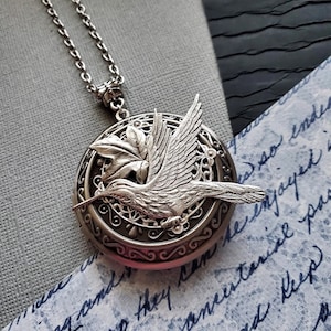 Puede incluir: Collar con dije de plata en forma de medallón adornado con un colibrí detallado y una hoja. El medallón está abierto y el interior es visible.