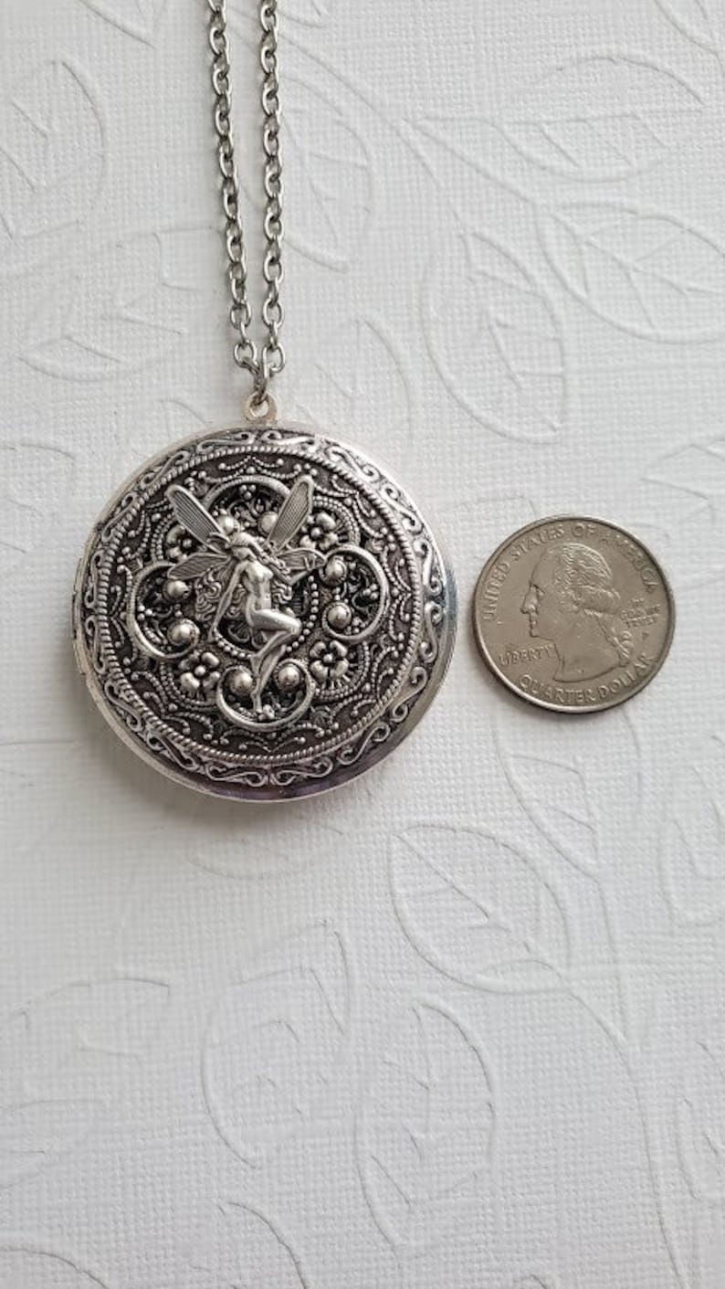 May include: Silver locket necklace with a detailed filigree design and a fairy charm. The locket is about the size of a quarter.