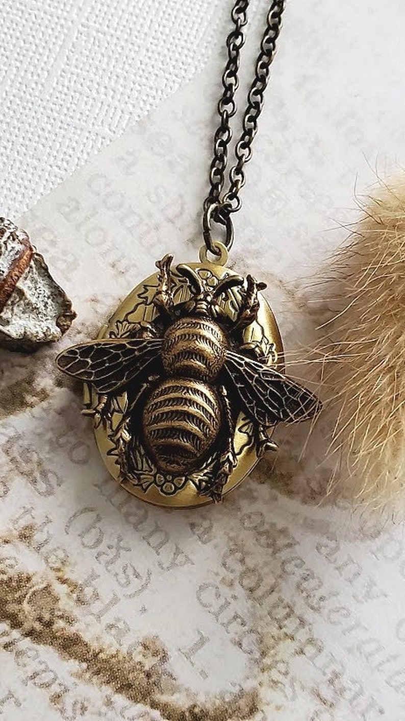 May include: A gold-toned locket necklace featuring a detailed bee design. The bee is surrounded by floral accents and has a textured, striped body.