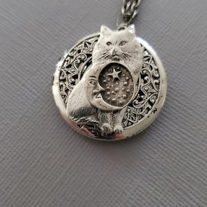 K&ouml;nnte beinhalten: Ein silberfarbener, runder Medaillon-Anh&auml;nger an einer passenden Kette. Das Medaillon zeigt ein erhabenes Relief einer Katze vor einem Halbmond- und Sternmotiv, eingebettet in einen Hintergrund aus filigranen, durchbrochenen Metalldetails.