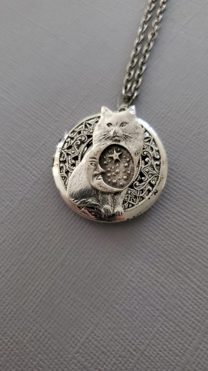 K&ouml;nnte beinhalten: Ein silberfarbener, runder Medaillon-Anh&auml;nger an einer passenden Kette. Das Medaillon zeigt ein erhabenes Relief einer Katze vor einem Halbmond- und Sternmotiv, eingebettet in einen Hintergrund aus filigranen, durchbrochenen Metalldetails.