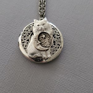 K&ouml;nnte beinhalten: Eine silberfarbene Medaillon-Halskette mit einem runden Anh&auml;nger, der einen filigranen Rand aufweist. In der Mitte des Anh&auml;ngers befindet sich eine erhabene Katzenfigur, die ein Motiv aus Mondsichel und Stern h&auml;lt. An einer passenden Kette.