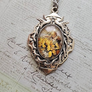 Puede incluir: Un collar de plata con un colgante de camafeo que presenta una ilustración vintage de dos hadas rodeadas de flores amarillas. El colgante está enmarcado por un diseño decorativo con dos pájaros en la parte superior.
