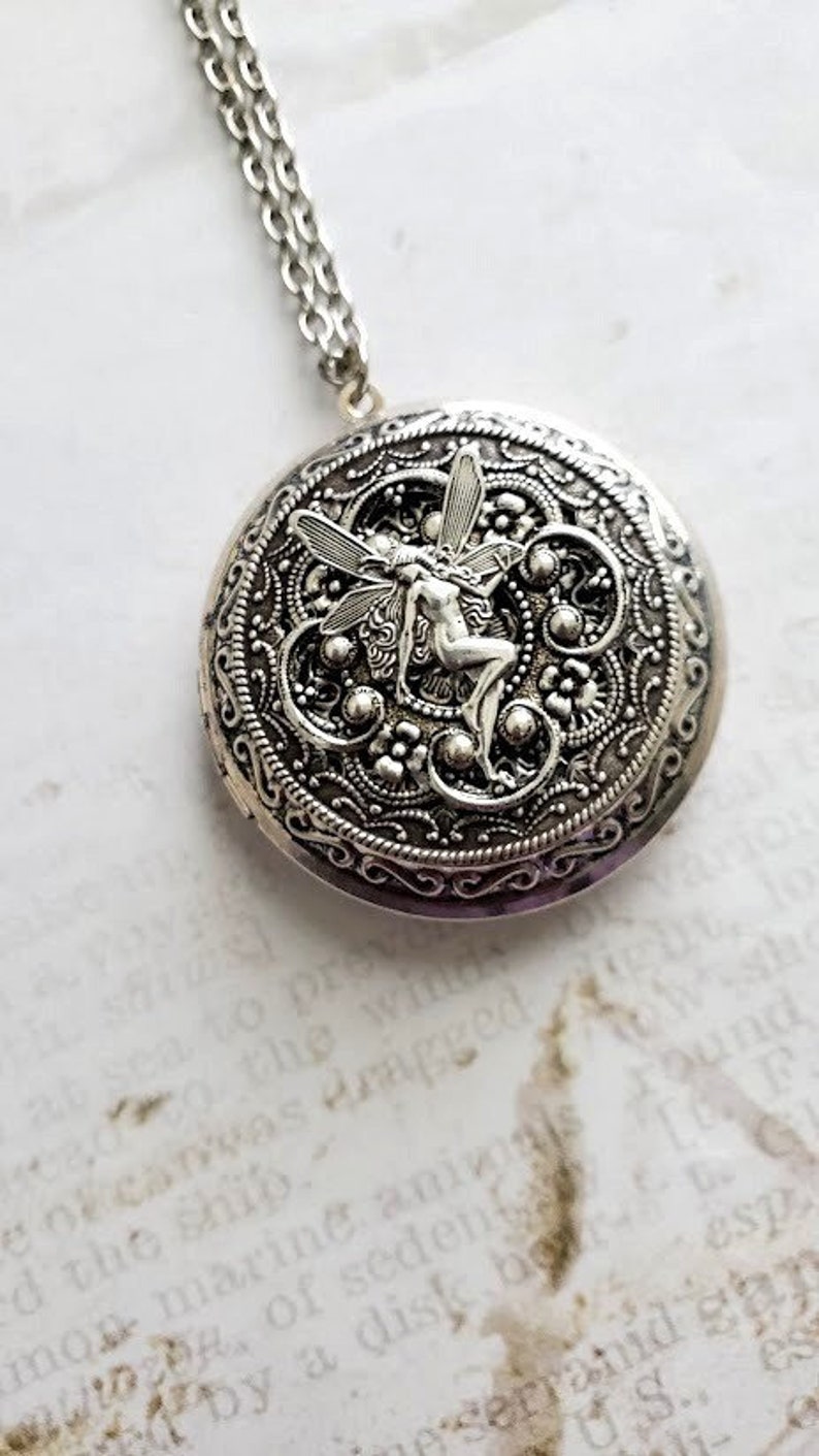 May include: Silver locket necklace with a detailed filigree design featuring a fairy with wings. The locket has a round shape and a silver chain.