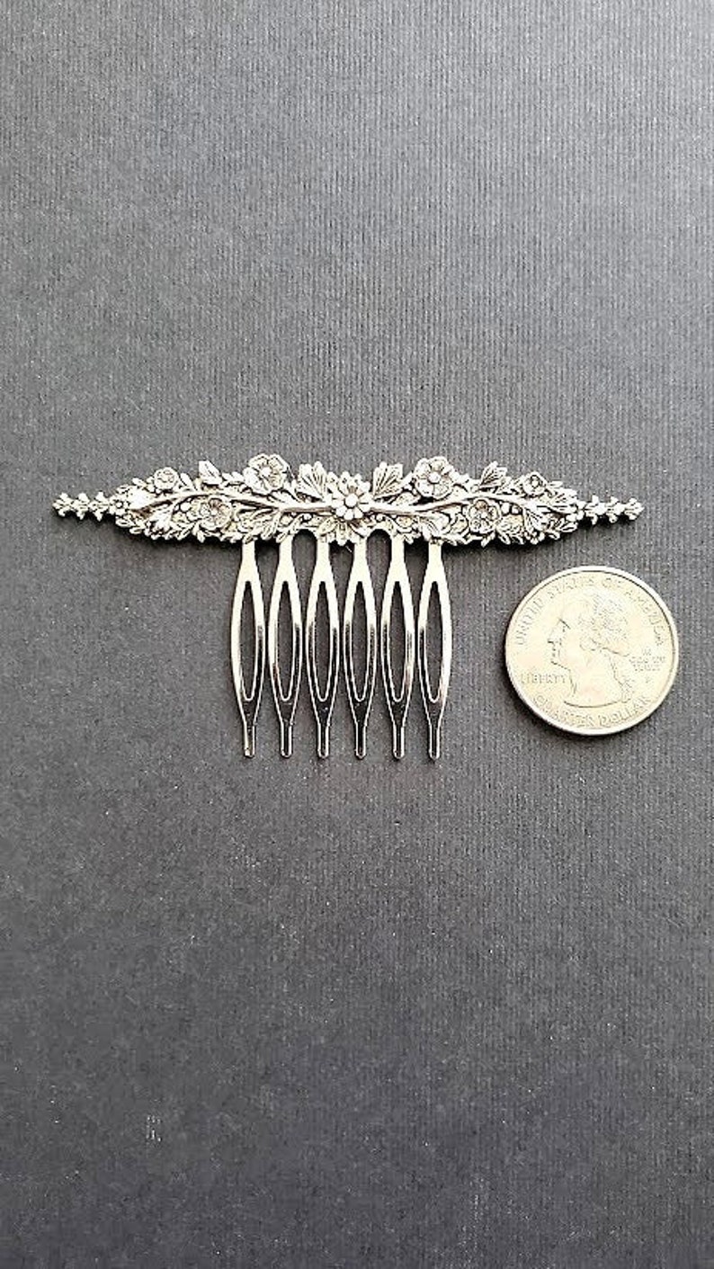 May include: A silver hair comb with a floral design. The comb has seven teeth and is about the size of a US quarter.