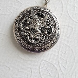 May include: Silver locket necklace with a detailed design featuring a fairy with wings. The locket is round and has a filigree pattern.