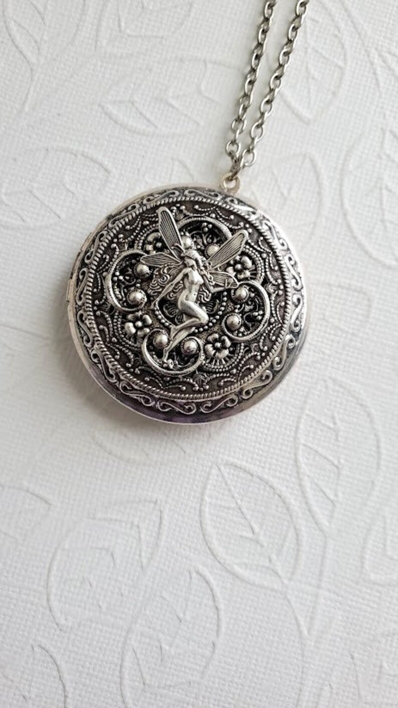 May include: Silver locket necklace with a detailed design featuring a fairy with wings. The locket is round and has a filigree pattern.