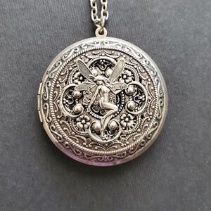 May include: Silver locket necklace with a detailed filigree design and a fairy figure in the center.