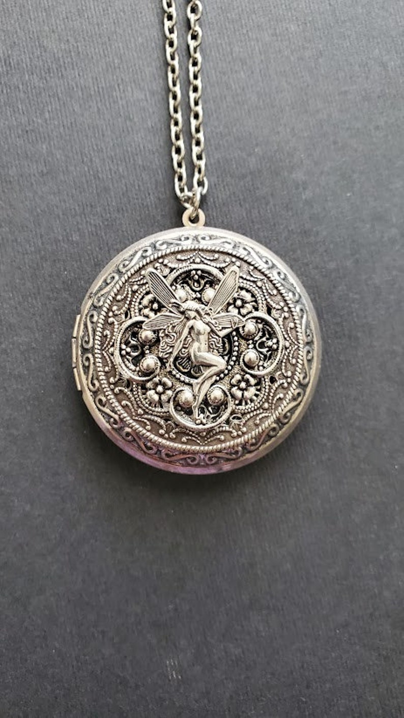 May include: Silver locket necklace with a detailed filigree design and a fairy figure in the center.