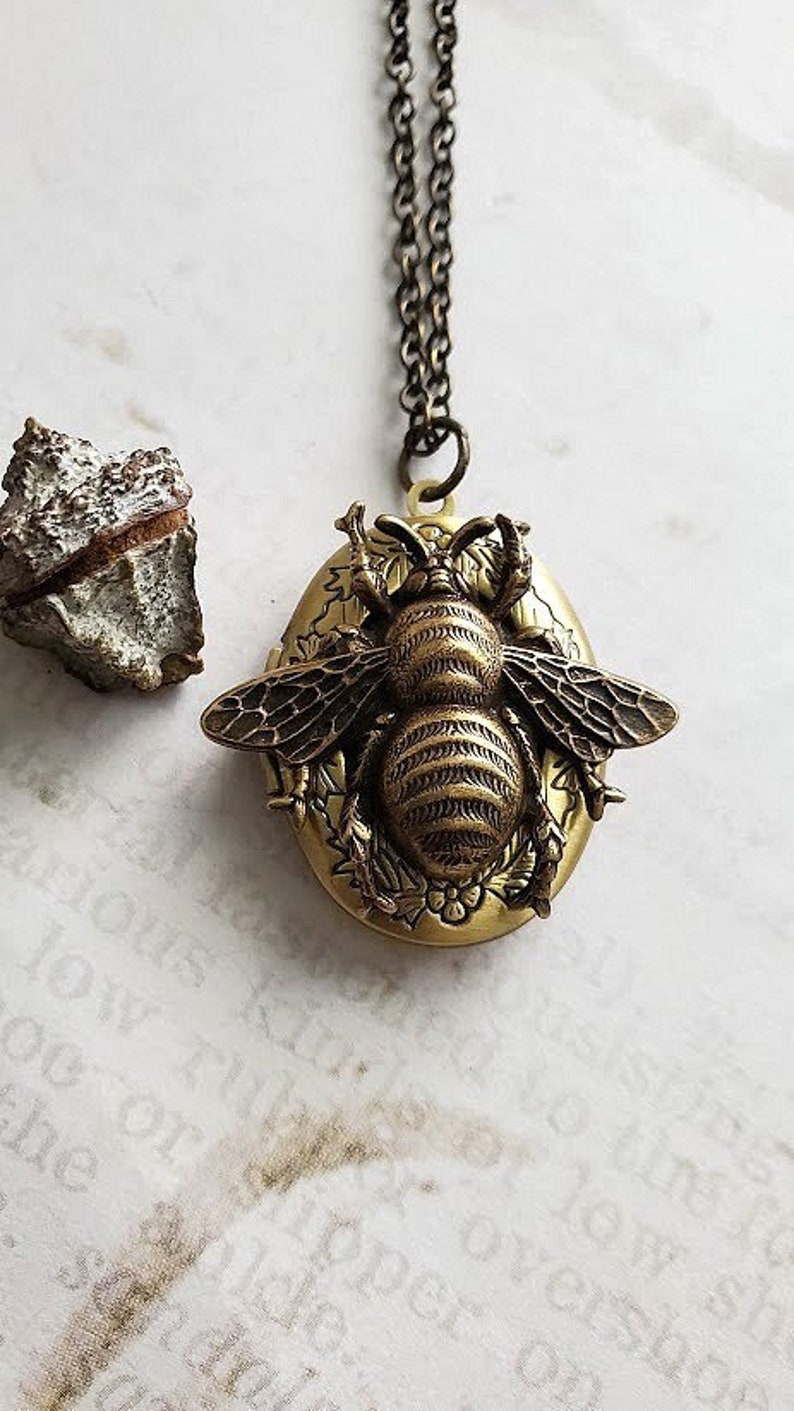 May include: A gold-toned bee locket necklace with a detailed bee design and floral accents. The locket is oval-shaped and hangs from a delicate chain.