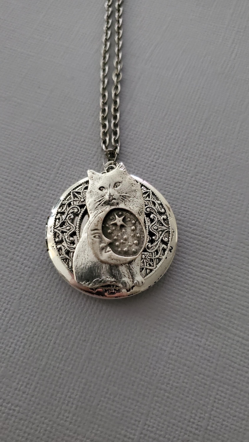 K&ouml;nnte beinhalten: Eine silberfarbene Medaillon-Halskette mit einem runden Anh&auml;nger, der einen filigranen Rand aufweist. In der Mitte befindet sich ein Relief einer Katze, die eine Mondsichel mit einem Stern und kleinen Punkten h&auml;lt, an einer passenden Gliederkette.