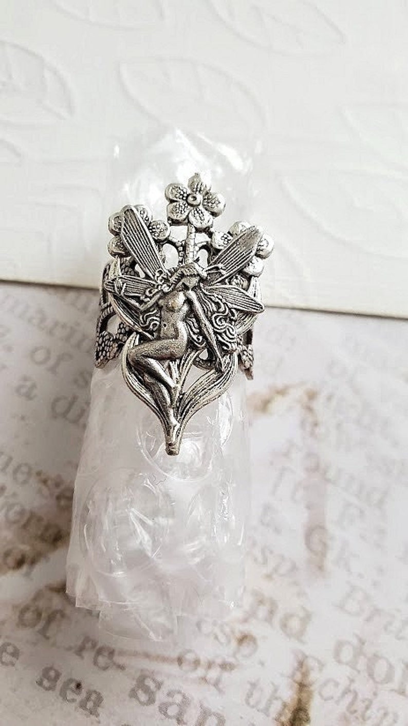May include: Silver ring with a detailed design of a fairy sitting on a flower with wings spread out.