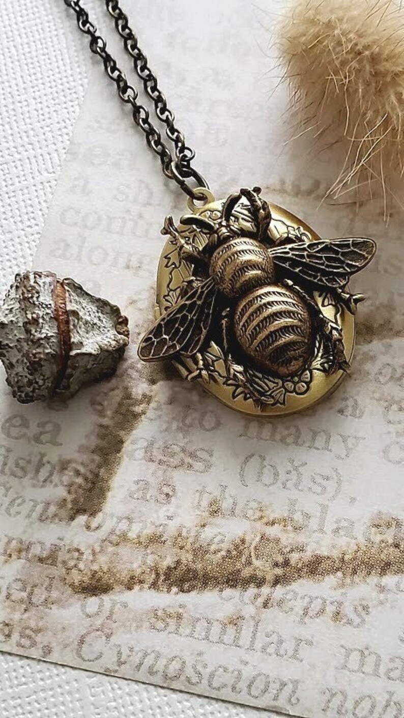 May include: A gold-toned locket necklace with a detailed bee design. The bee is surrounded by floral accents and has a textured, striped body. The necklace has a black chain.