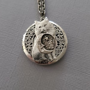 K&ouml;nnte beinhalten: Ein silberfarbener, runder Medaillon-Anh&auml;nger an einer passenden Kette. Das Medaillon zeigt ein erhabenes Relief einer Katze, die eine Mondsichel mit einem Stern und kleinen Punkten h&auml;lt. Der Hintergrund hat ein filigranes, durchbrochenes Muster.