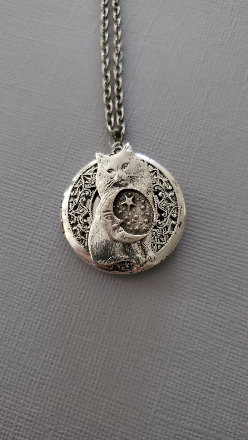 K&ouml;nnte beinhalten: Ein silberfarbener, runder Medaillon-Anh&auml;nger an einer passenden Kette. Das Medaillon zeigt ein erhabenes Relief einer Katze, die eine Mondsichel mit einem Stern und kleinen Punkten h&auml;lt. Der Hintergrund hat ein filigranes, durchbrochenes Muster.