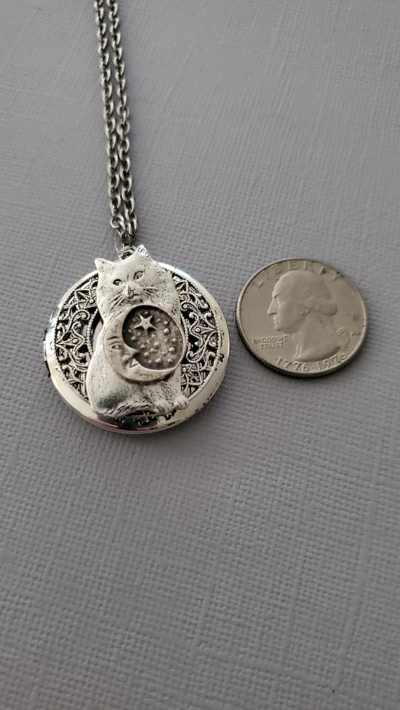 K&ouml;nnte beinhalten: Eine silberfarbene, kreisf&ouml;rmige Medaillon-Halskette mit einer Katze, die ein Mondsichel- und Sternmotiv h&auml;lt. Das Medaillon hat einen filigranen Rand und h&auml;ngt an einer passenden Kette, abgebildet neben einer 25-Cent-M&uuml;nze zum Gr&ouml;&szlig;envergleich.