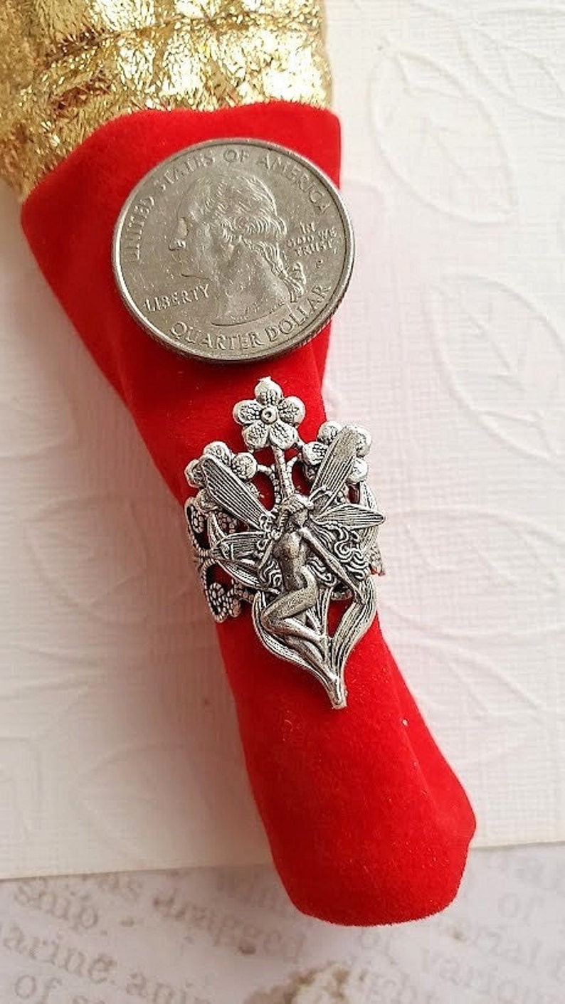 May include: Silver ring with a fairy design. The fairy is sitting on a flower with wings spread out. The ring is on a red velvet background.