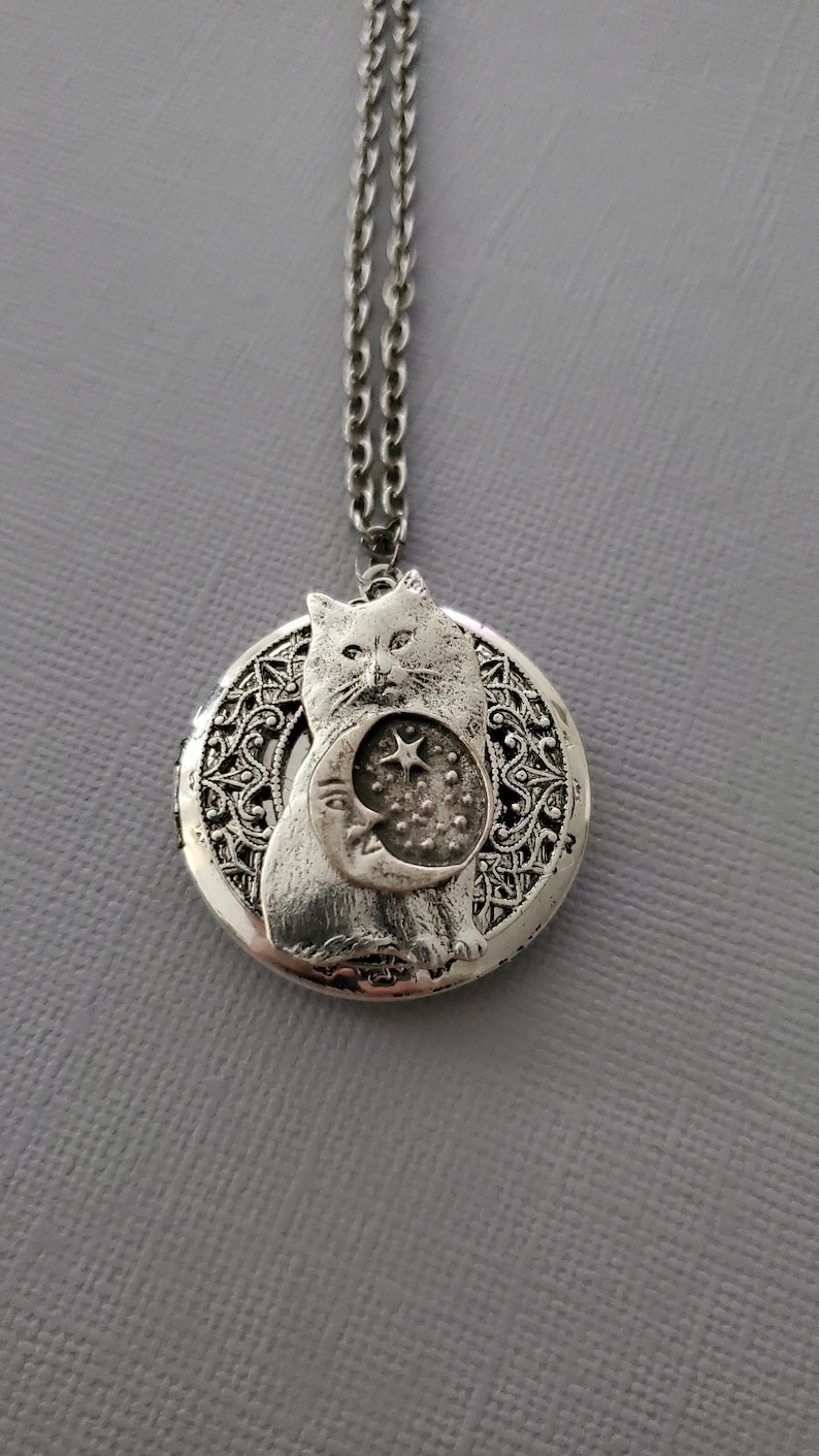 K&ouml;nnte beinhalten: Ein silberfarbener, runder Medaillon-Anh&auml;nger an einer passenden Kette. Das Medaillon zeigt ein erhabenes Relief einer Katze, die eine Mondsichel mit einem Stern und kleinen Punkten h&auml;lt. Der Hintergrund hat ein komplexes Filigranmuster.