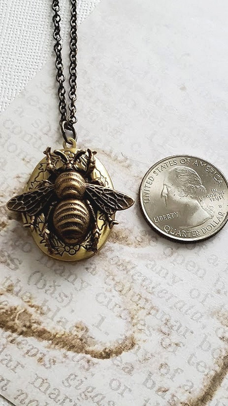 May include: A gold tone locket necklace featuring a detailed bee design. The locket is oval shaped and has a floral design around the bee. The necklace is on a black chain.