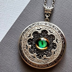 May include: Silver locket necklace with an ornate design and a green eye in the center.