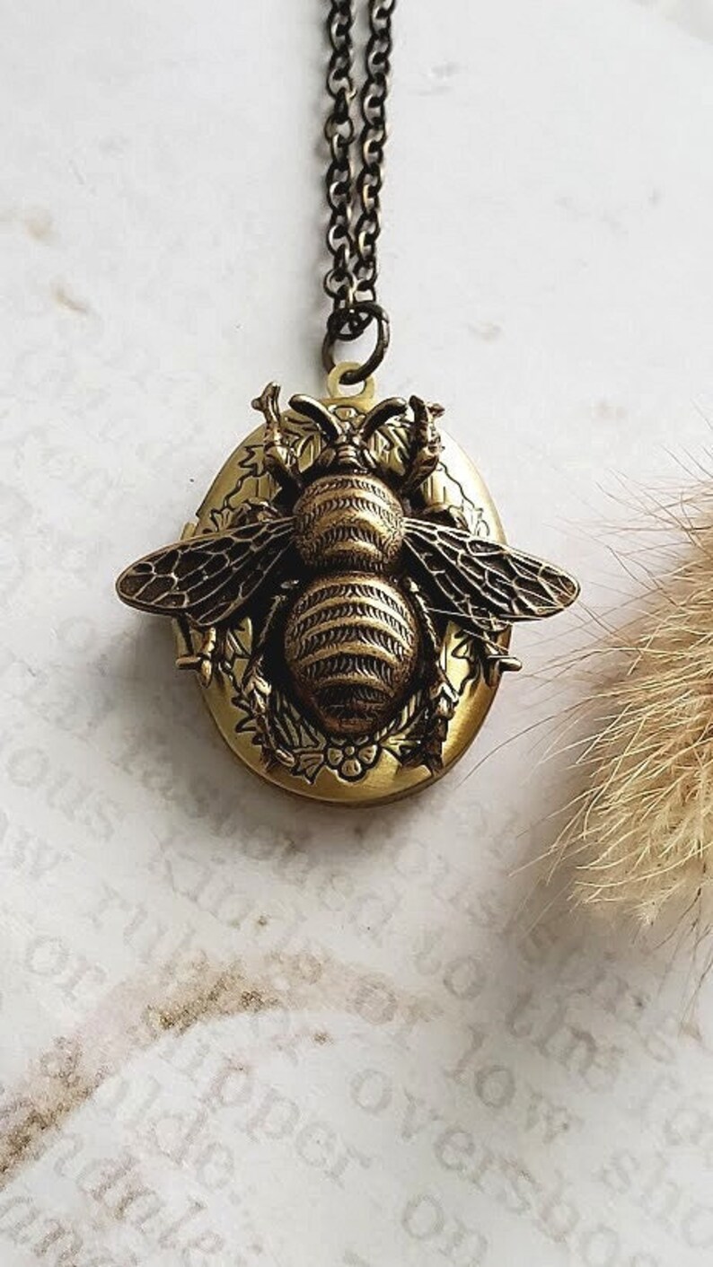 May include: A gold tone locket necklace featuring a detailed bee design. The bee is surrounded by floral accents and is set against a gold tone oval background.